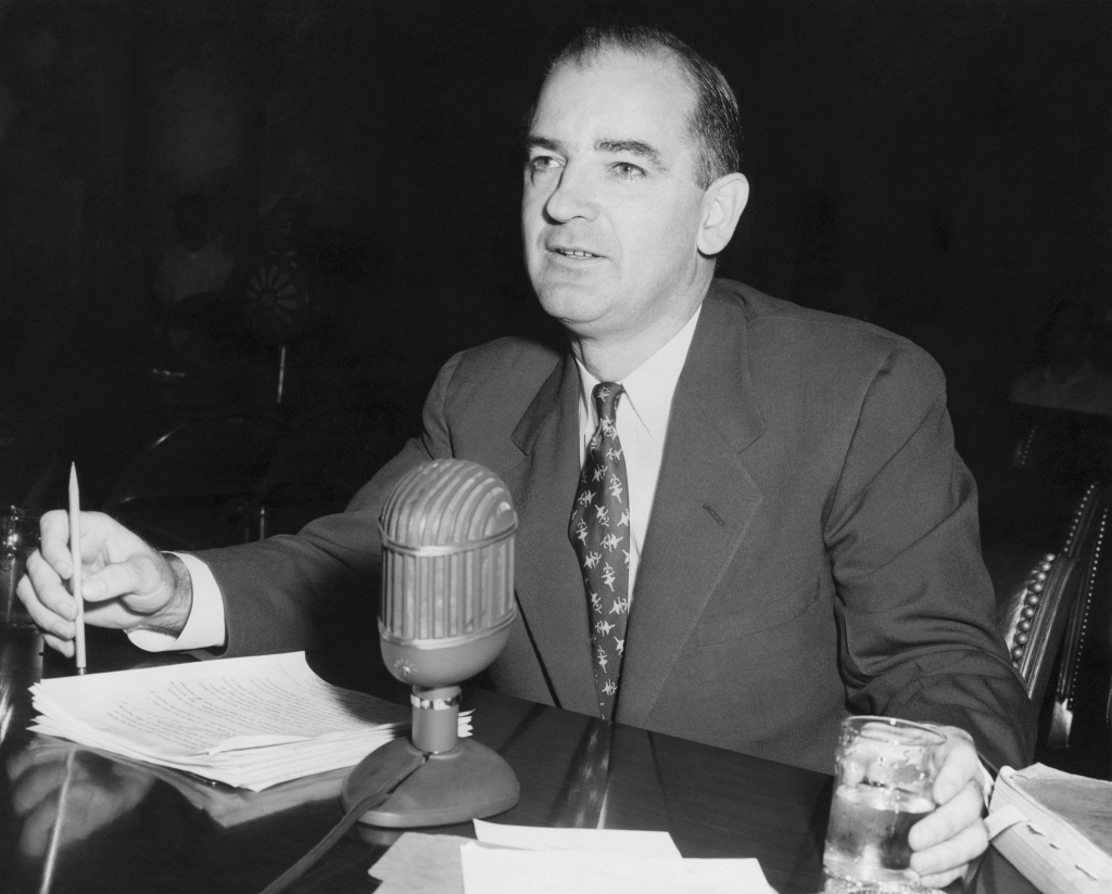 Senator Joseph McCarthy holding a pen and speaking at a microphone during a hearing